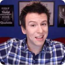 Man with short dark hair wearing a blue and white plaid shirt, smiling and speaking in front of a blurred background with framed posters.